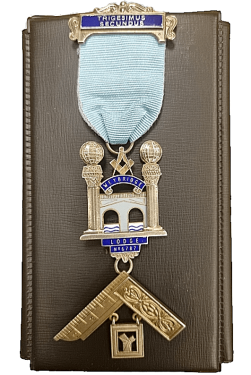 Freemasons in Guildford Surrey. A Past Masters Jewel depicting the bridge over the River Wey at Weybridge between the two columns from the entrance of King Solomon temple with a builders square set below. The top ribbon is adorned with a plate inscribed in latin 'Trigesimus Secundus' or thirty second. The jewel is cast from 18ct gold and enamelled with a light blue ribbon.